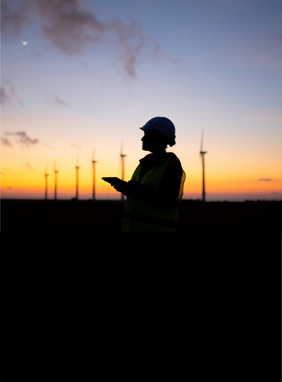 Silhouette of person with construction hat against a sunrise horizon and wind turbines in the background
